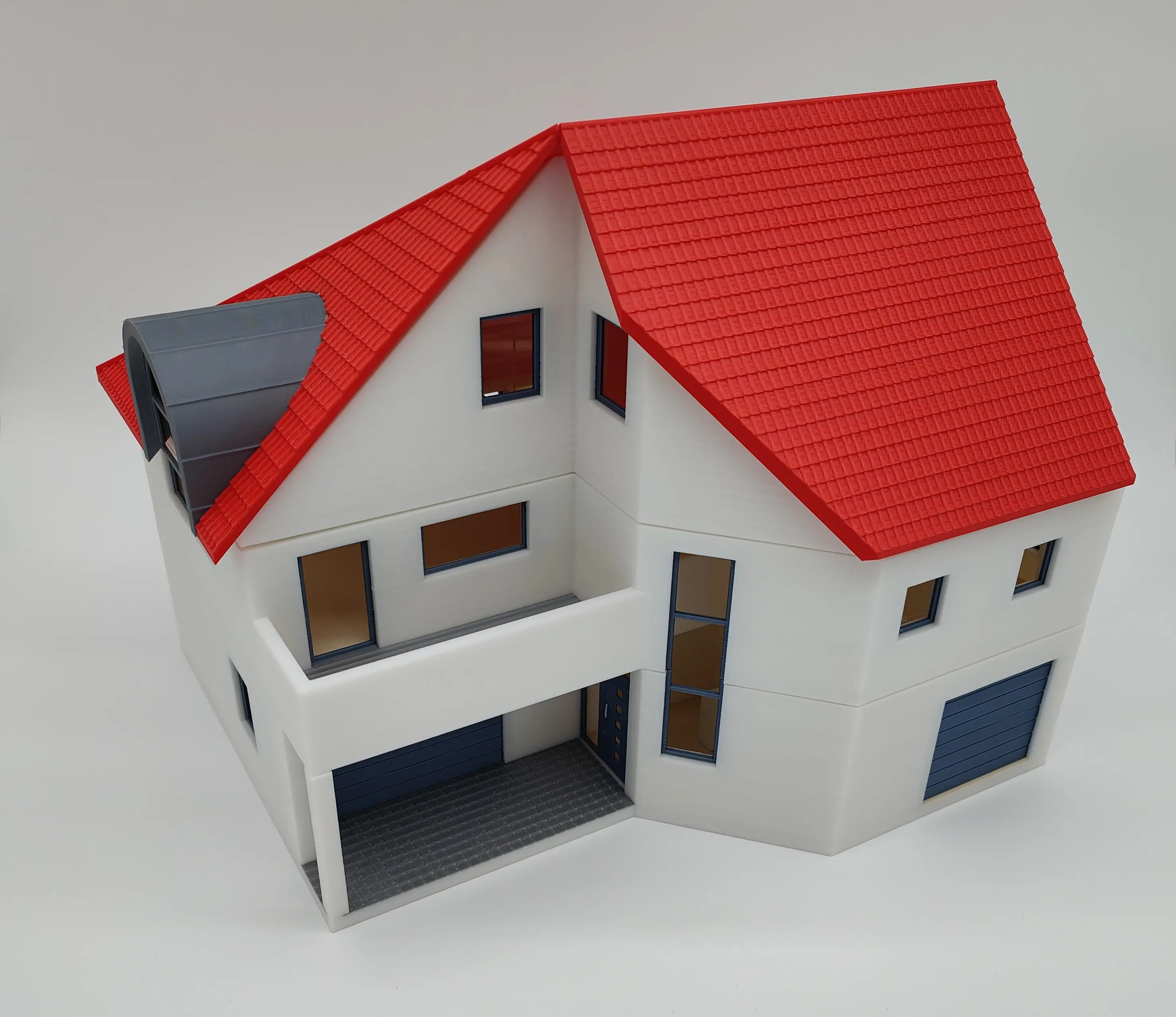 Detailliertes 3D-gedrucktes Modell eines Hauses aus der Vorderperspektive, mit weißem Mauerwerk, rotem Ziegeldach und schwarzen Solarpanelen. Die Fensterrahmen, Haustür und das Garagentor sind grau, vor einem weißen Hintergrund.