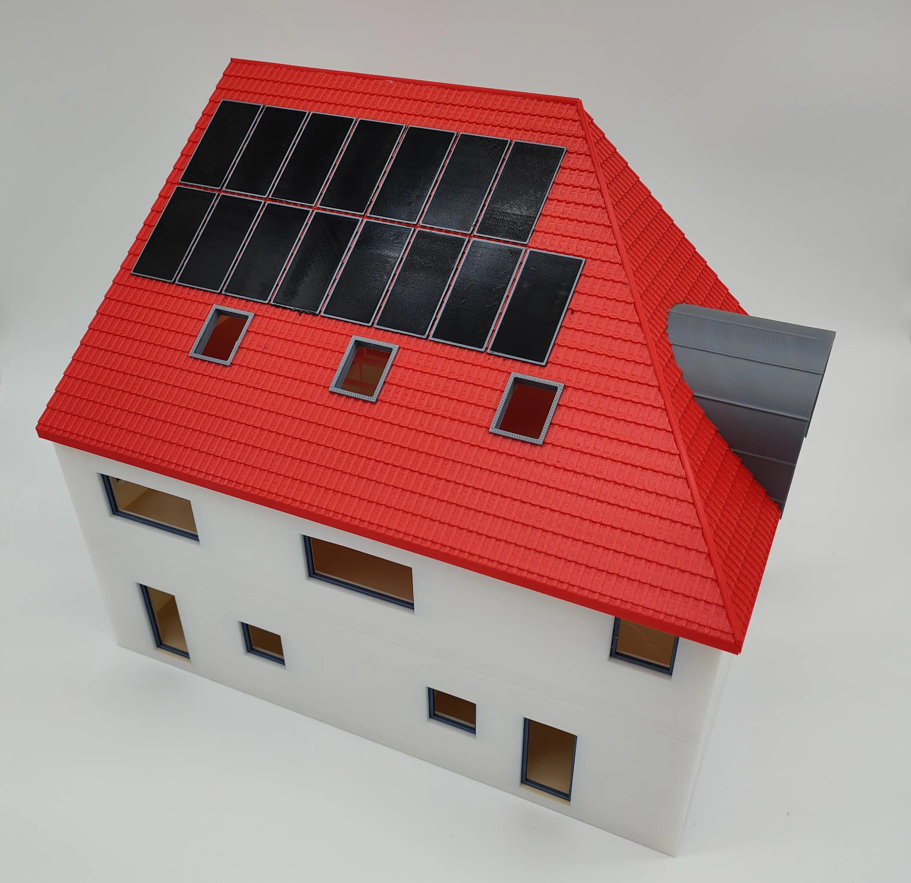 Großes, detailliertes 3D-gedrucktes Modell eines Hauses mit weißen Wänden, rotem Ziegeldach und schwarzen Solarpanelen. Die Fensterrahmen und die Gaube sind grau, vor einem weißen Hintergrund.