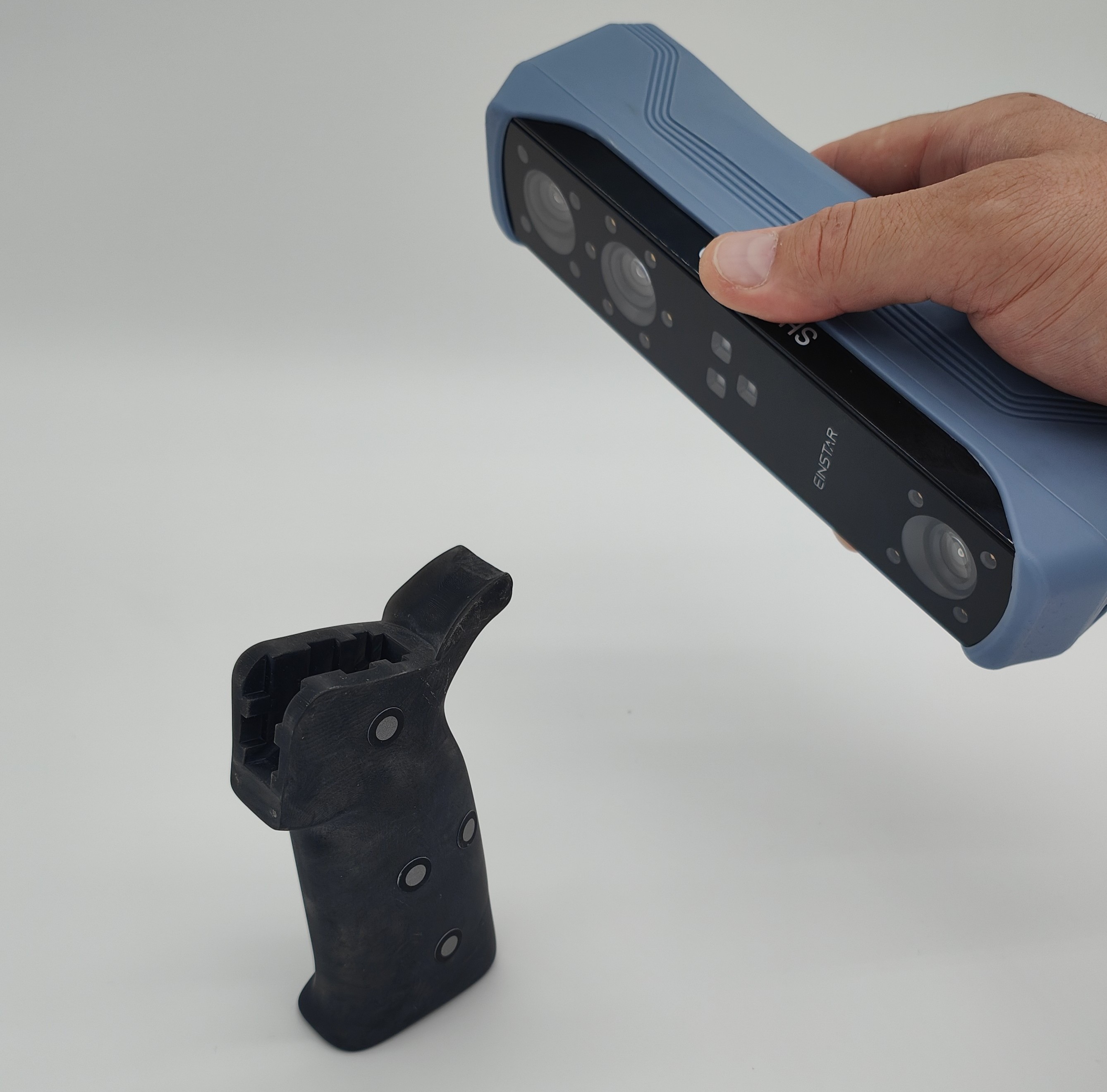 Eine Hand hält einen 3D-Scanner, der ein schwarzes Bauteil mit reflektierenden Scan-Markern erfasst. Der Hintergrund ist weiß, wodurch der Fokus auf dem präzisen Scannen des Bauteils liegt.