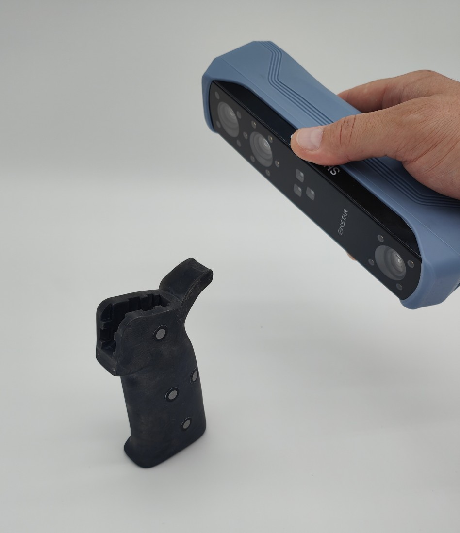 Eine Hand hält einen 3D-Scanner, der ein schwarzes Bauteil mit reflektierenden Scan-Markern erfasst. Der Hintergrund ist weiß, wodurch der Fokus auf dem präzisen Scannen des Bauteils liegt.