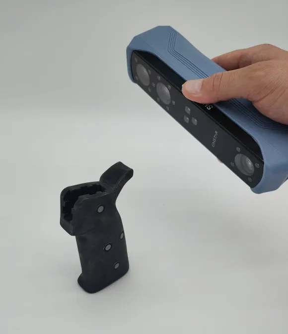 Eine Hand hält einen 3D-Scanner, der ein schwarzes Bauteil mit reflektierenden Scan-Markern erfasst. Der Hintergrund ist weiß, wodurch der Fokus auf dem präzisen Scannen des Bauteils liegt.
